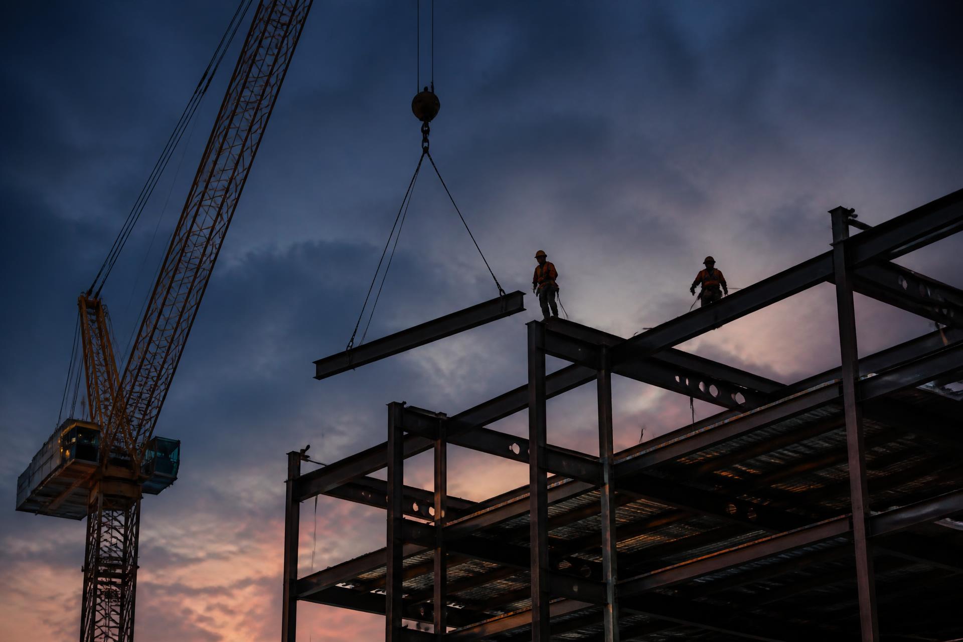Steel frame under erection with a tower crane lifting a beam at dusk